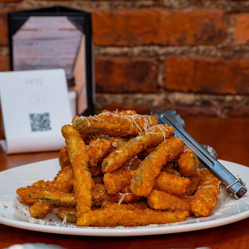 Crispy fried zucchini sticks, sprinkled with Parmesan cheese, on a white plate with tongs.