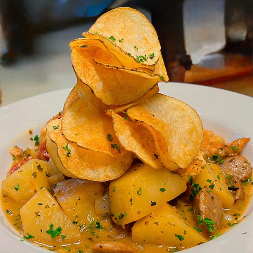 A white bowl filled with chunky potatoes, sausage, and a creamy sauce, topped with crispy potato chips.