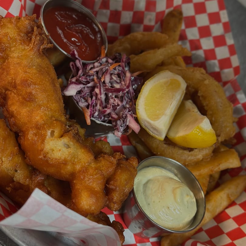 Golden-brown fish and chips with coleslaw, lemon wedges, ketchup, and tartar sauce in a red checkered basket.