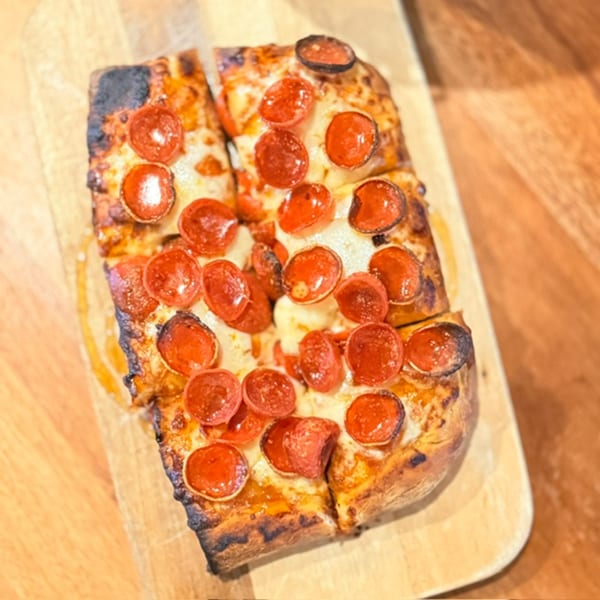 Pepperoni pizza with melted cheese and charred crust served on a wooden board, accompanied by a small cup of dipping sauce.