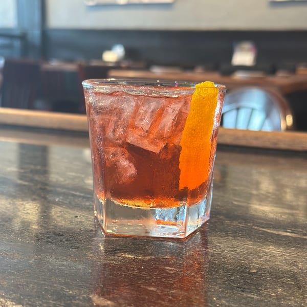 A vibrant red cocktail with ice and an orange peel garnish sits on a dark bar top.