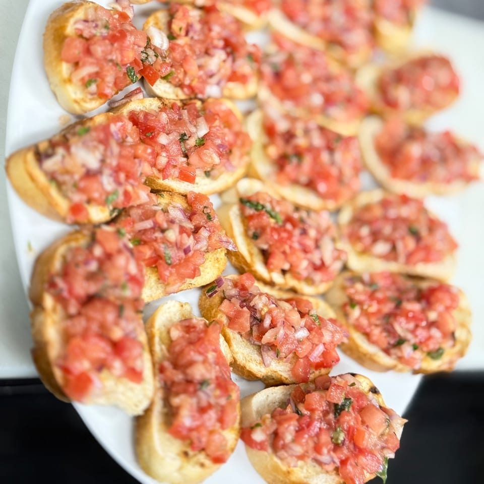 Close-up of a white platter filled with numerous bruschetta appetizers topped with diced tomatoes and herbs.