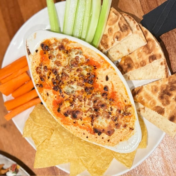 Baked cheese and bean dip garnished with tomatoes, served with tortilla chips, carrots, celery, and toasted flatbread.