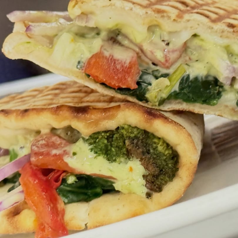 A grilled panini sandwich, cut in half, revealing broccoli, red peppers, spinach, and creamy sauce.