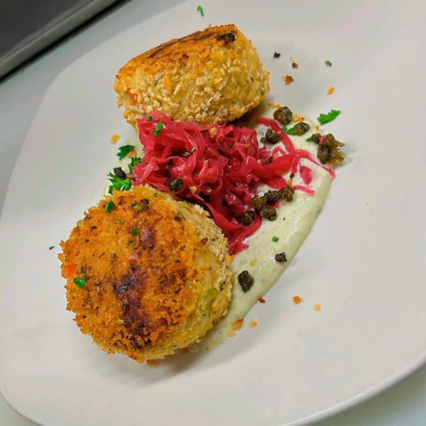Two golden-brown crab cakes with pickled red onions, capers, and a creamy sauce on a white plate.