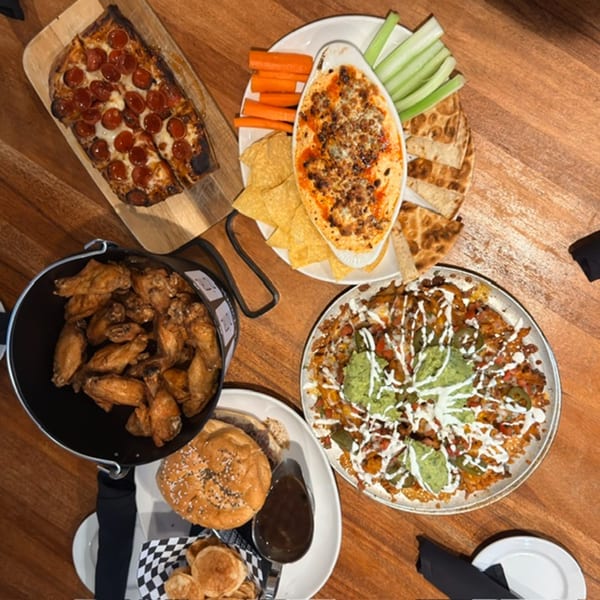 Overhead spread of pepperoni pizza, chicken wings, loaded nachos, a burger, flatbread, dip, and fresh vegetable sticks on a wooden table.