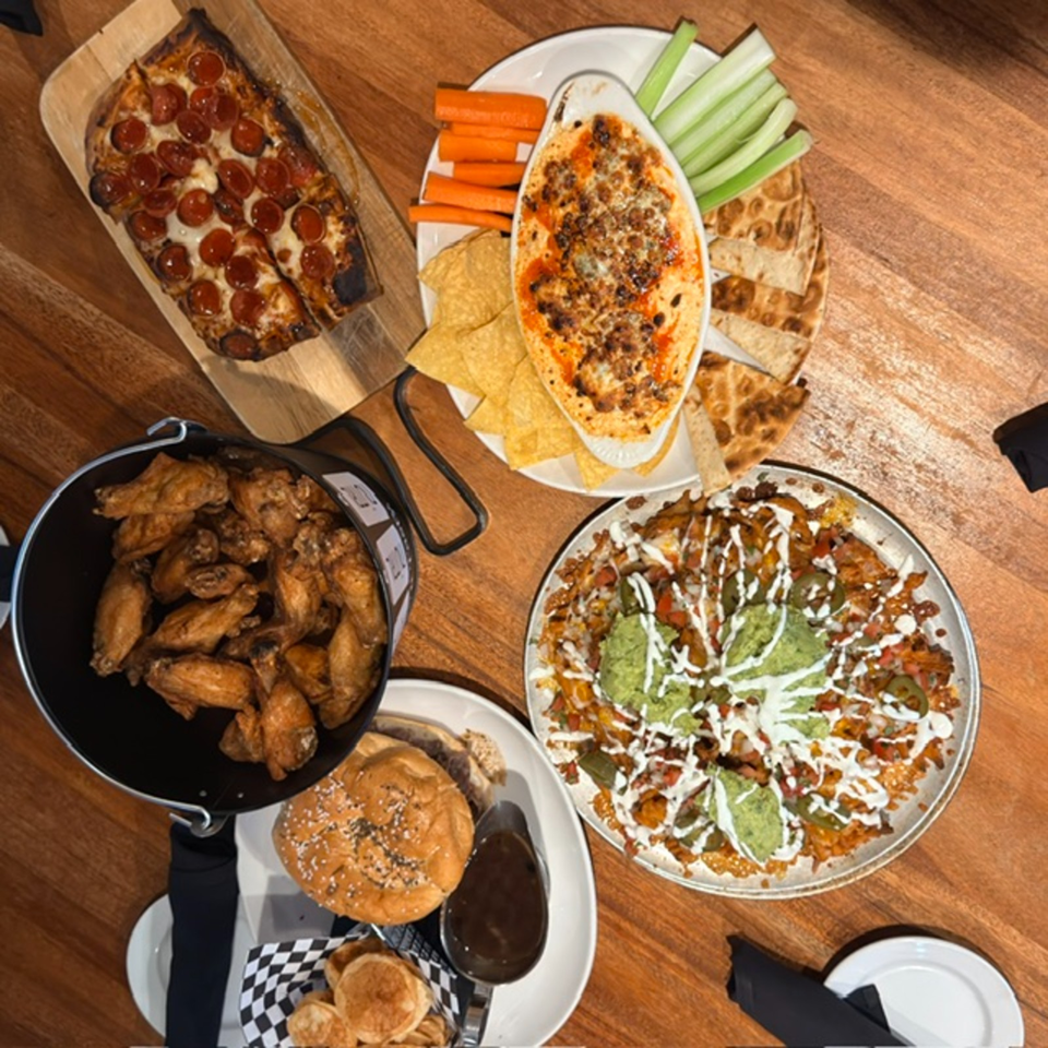 Overhead spread of pepperoni pizza, chicken wings, loaded nachos, a burger, flatbread, dip, and fresh vegetable sticks on a wooden table.